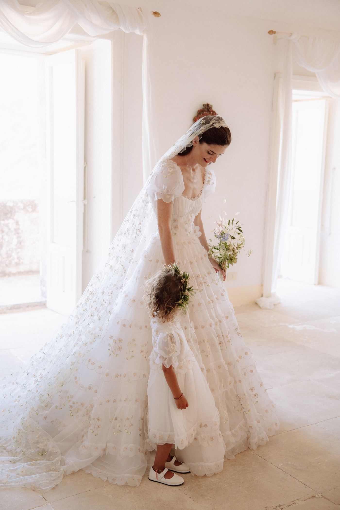 Bride in an embroidered organza gown and floral tulle veil, with a flower girl, in a sunlit white room