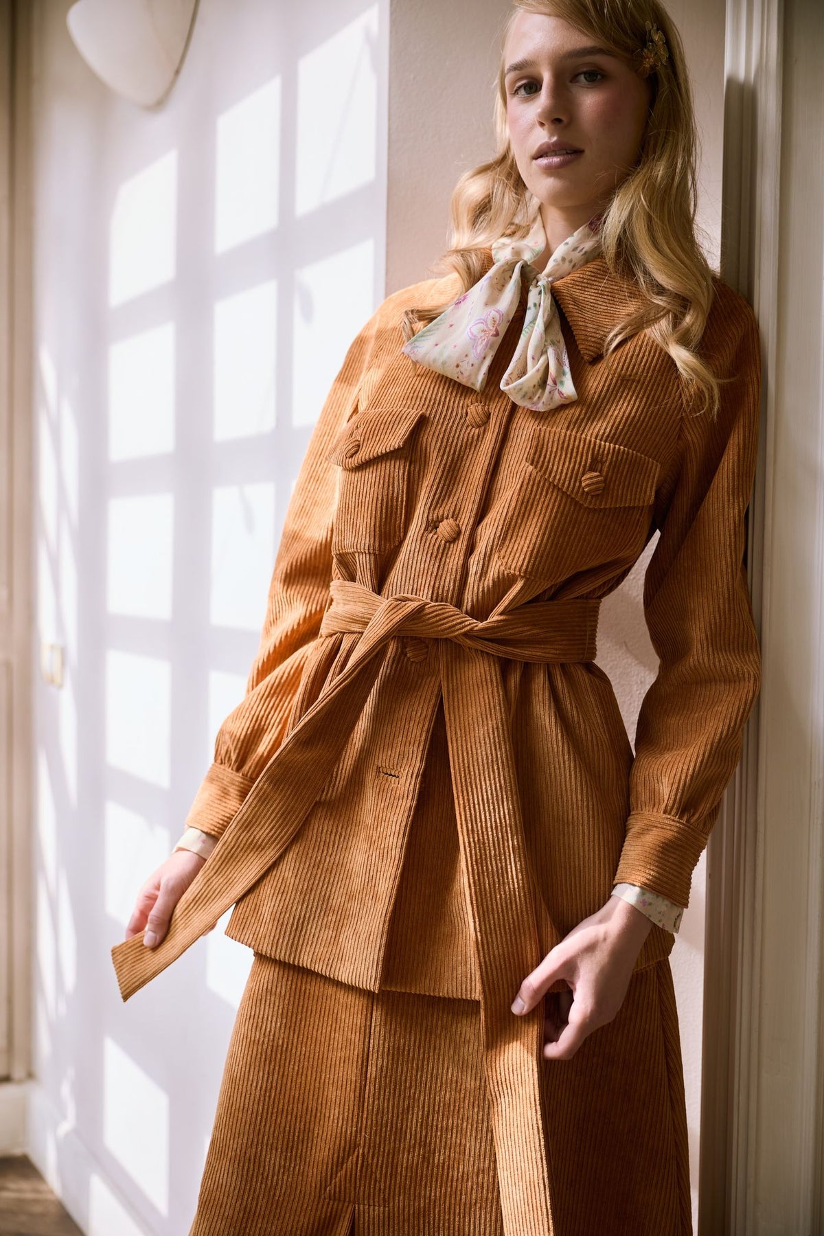 Model in a caramel corduroy jacket and skirt with a knotted belt and floral scarf